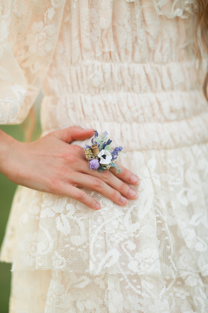 Close-up of a hand holding a small floral purple ring against a lace dress background
