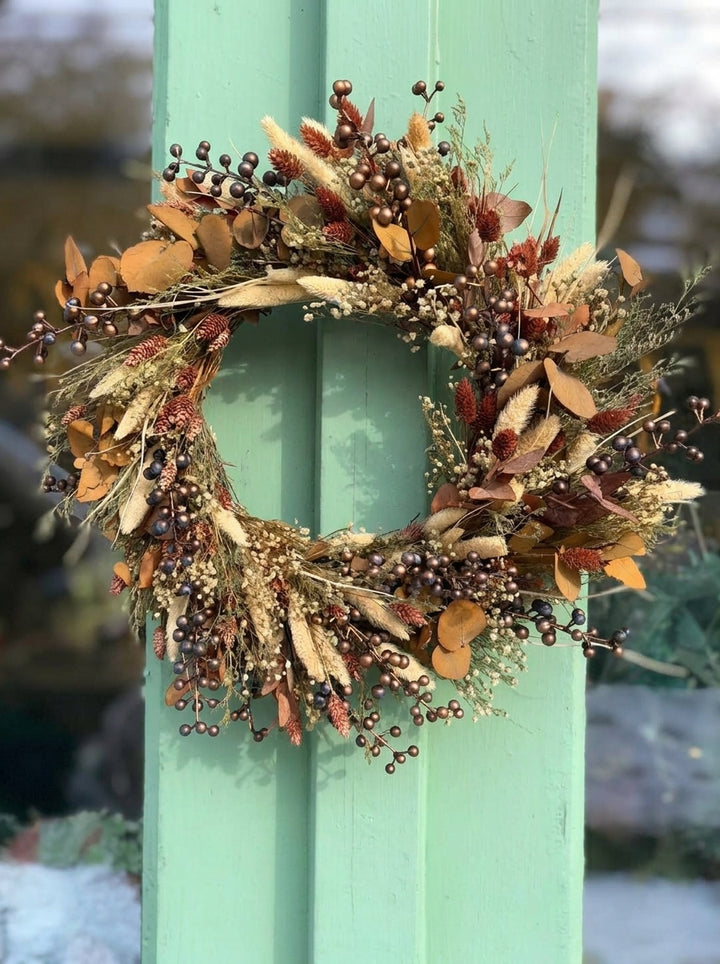 An autumn-themed decorative wreath with dried grasses, small brown berries, and tan leaves hanging on a mint green painted wooden post.
