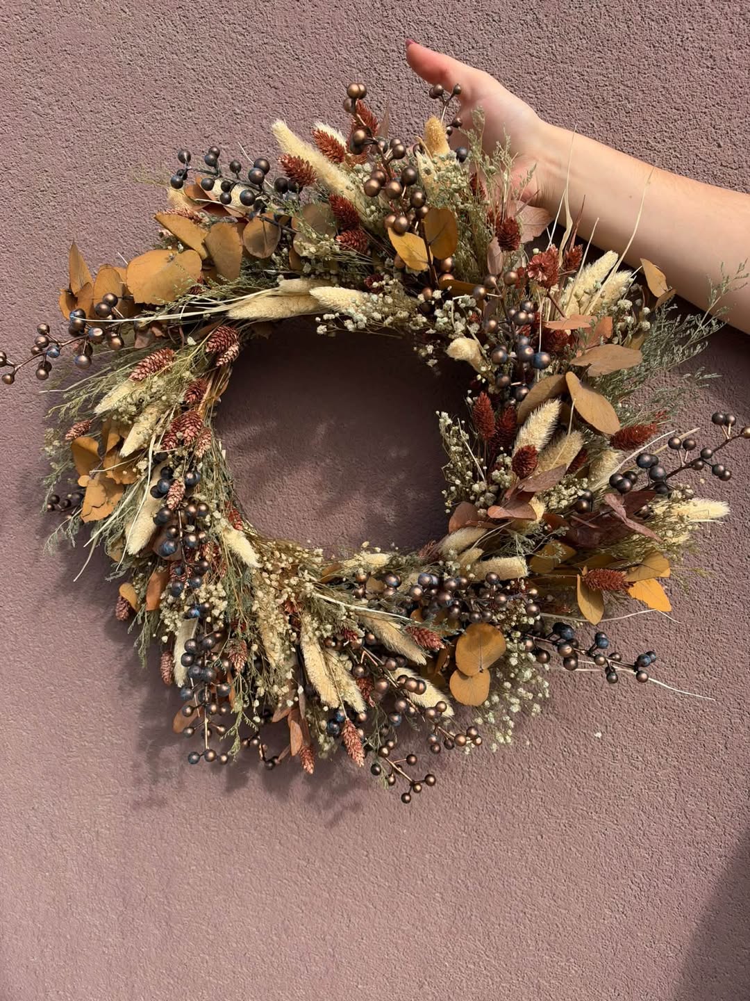 A dried floral wreath with beige grasses, brown leaves, and small dark berries, held by a person's hand against a dusty mauve wall.