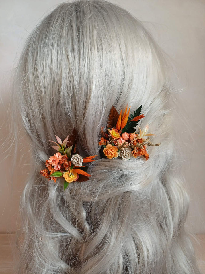 A close-up of long, wavy silver-gray hair from the back, decorated with two small floral hair clips in orange, beige, and green.