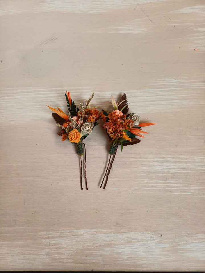 Two small floral bobby pins with orange, brown, and cream flowers, attached to dark hairpins, displayed on a light beige wood background.