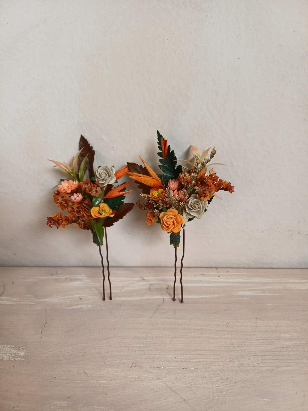 Two small floral hair accessories with orange, brown, beige, and green botanical elements on thin metal backs, displayed against a light textured wall and pale wood surface.