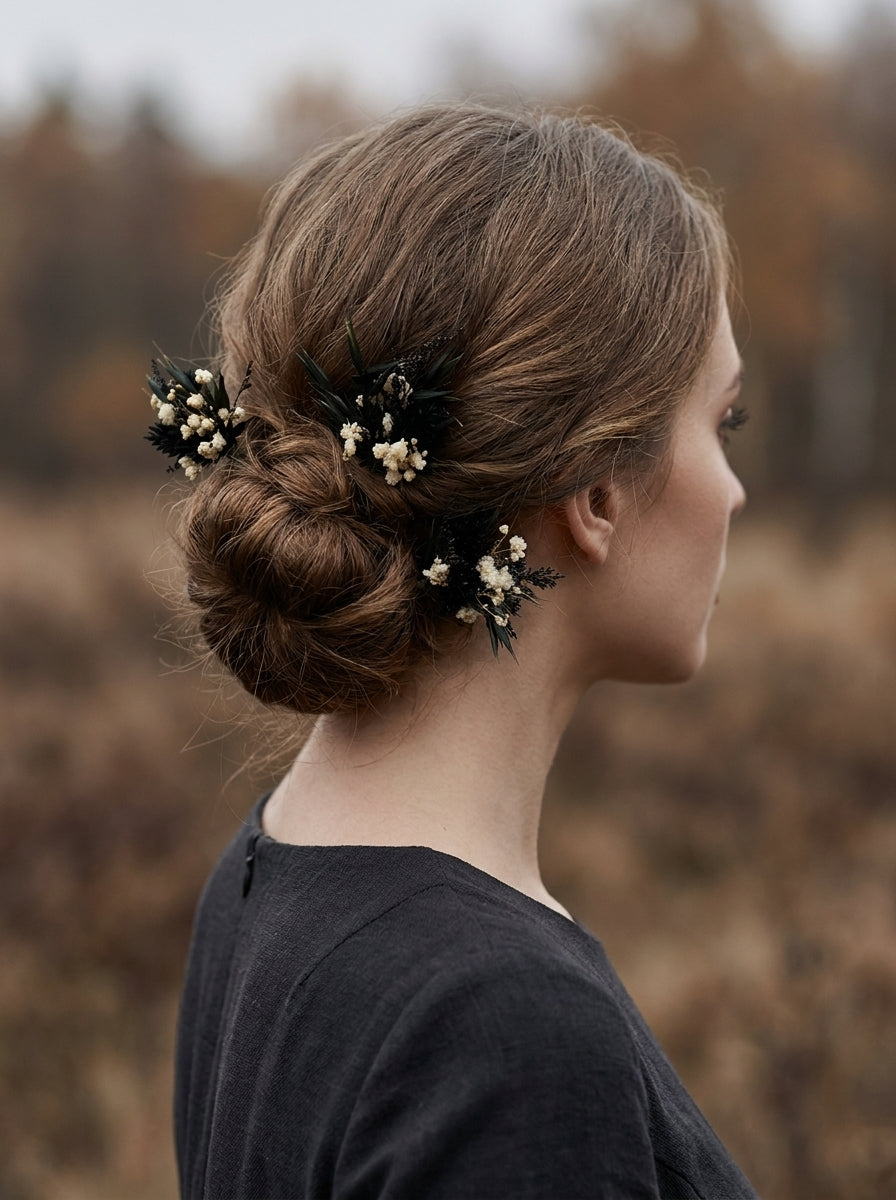 A woman in profile wearing a dark gray top, showcasing a styled brown updo with delicate black and cream floral hair accessories, set against a blurred autumnal field background.