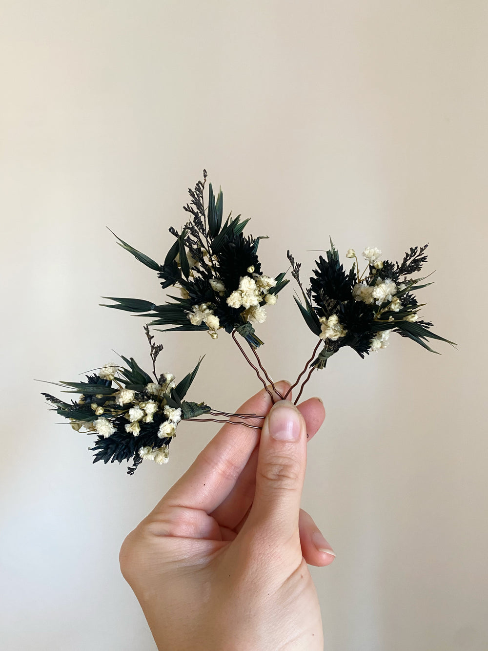 A hand holding a dried floral arrangement featuring dark green leaves and small cream baby's breath flowers against a plain beige background.