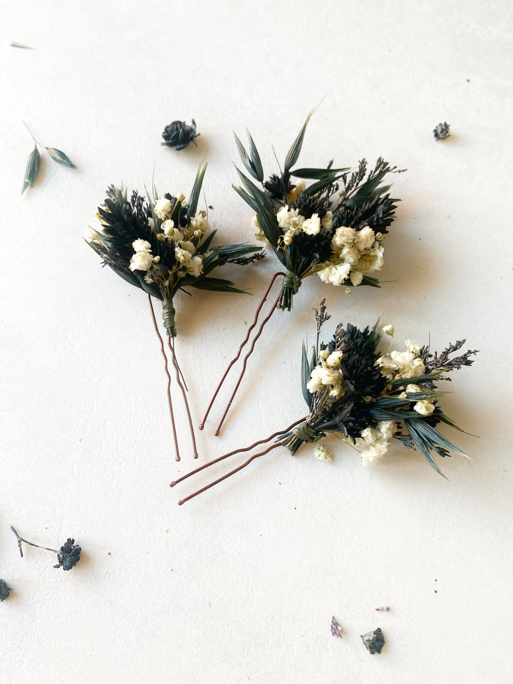 Three dried floral boutonnieres with white baby's breath, dark green leaves, and blue-green accents, attached to brown hair pins and arranged on a light beige textured background.