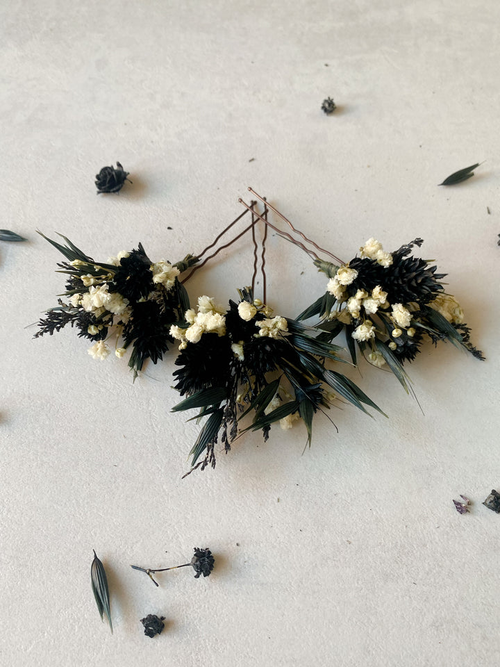 Three dried floral hair accessories with black seed pods, white baby's breath, and green leaves, arranged on a light beige textured surface.