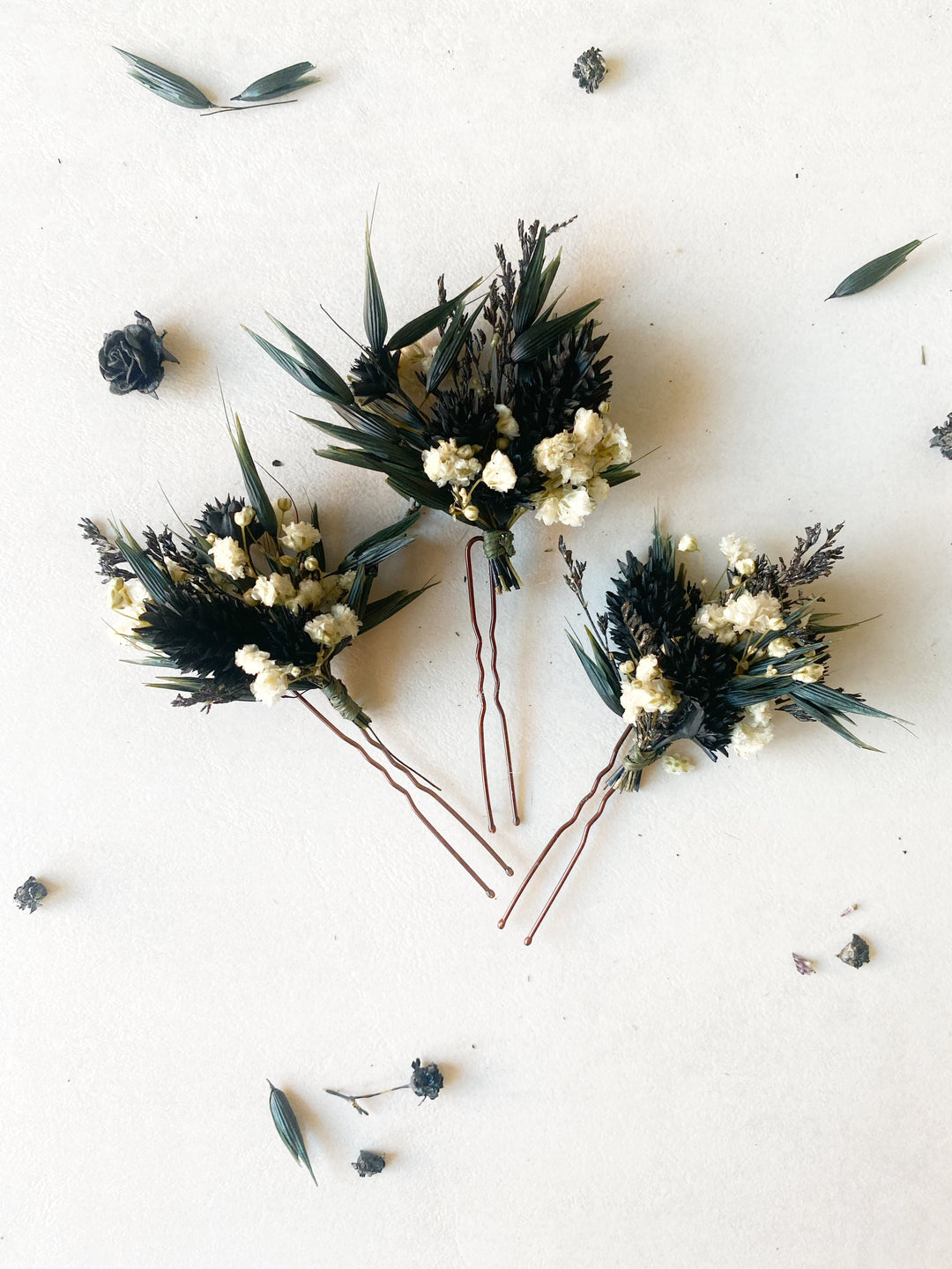 Three dried floral boutonniere-style hairpins with dark green leaves, cream blossoms, and brown stems, arranged on a light beige background with scattered dried botanicals.