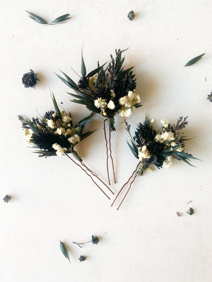 Three dried floral boutonniere-style hairpins with dark green leaves, cream blossoms, and brown stems, arranged on a light beige background with scattered dried botanicals.