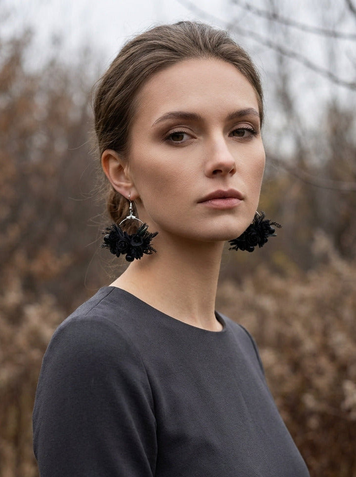 A woman with her hair pulled back wears large black flower-shaped dangle earrings and a dark charcoal long-sleeve top, standing outdoors against a blurred background of dry grass and trees.