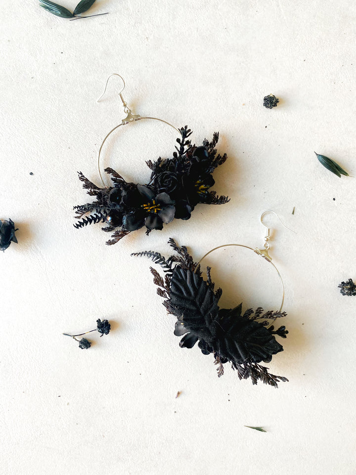 A pair of large gold-tone hoop earrings adorned with black dried flowers, pinecones, and small black berries, arranged on a textured off-white surface with scattered botanical elements.