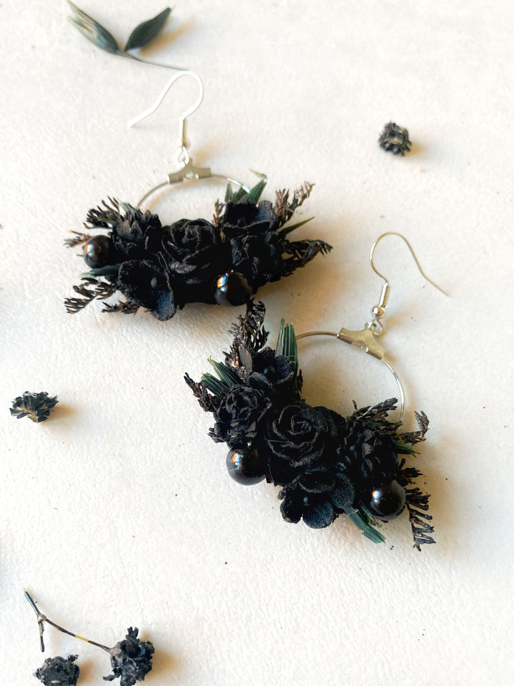 Black floral hoop earrings with black roses, pinecones, and green leaves, displayed on a textured off-white background with scattered dried botanicals.