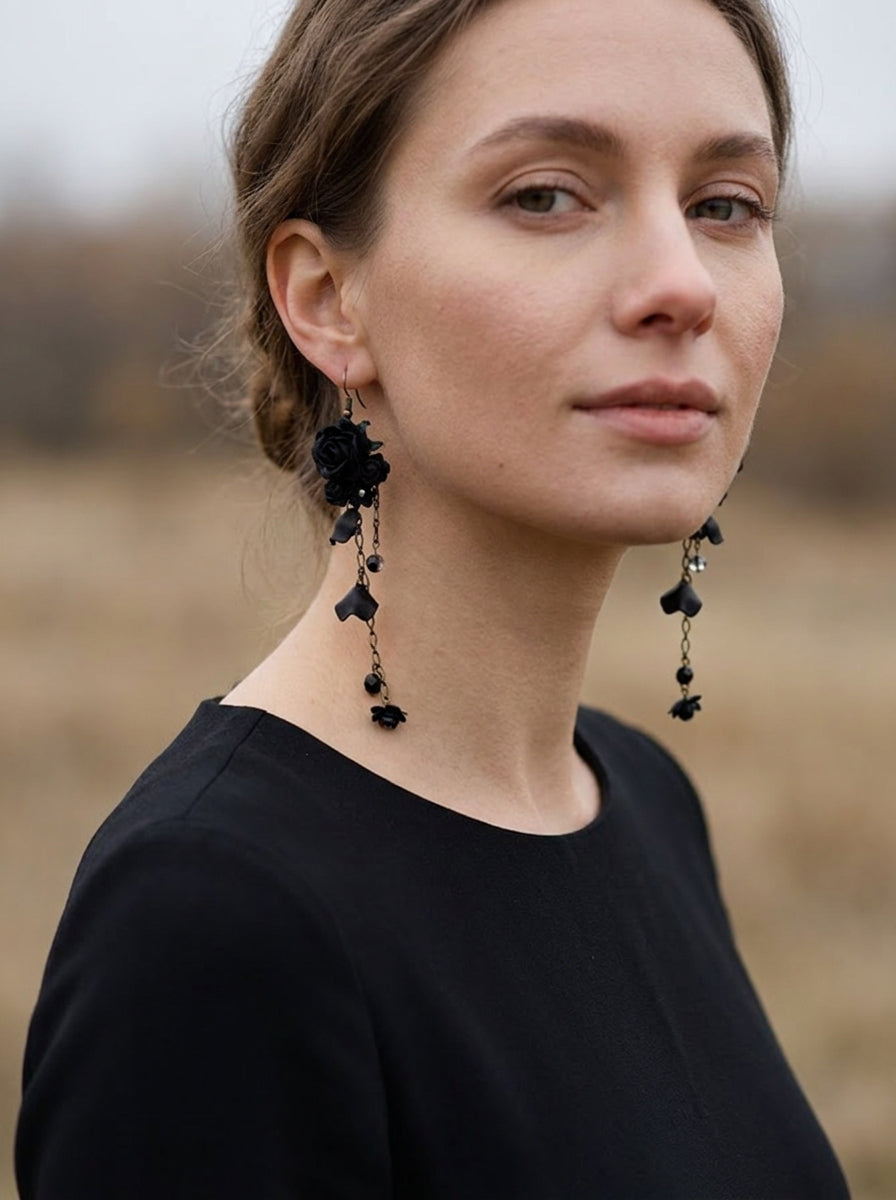 A woman wearing long black rose dangle earrings with small black flower charms and dark beads, photographed outdoors against a softly blurred tan field background.