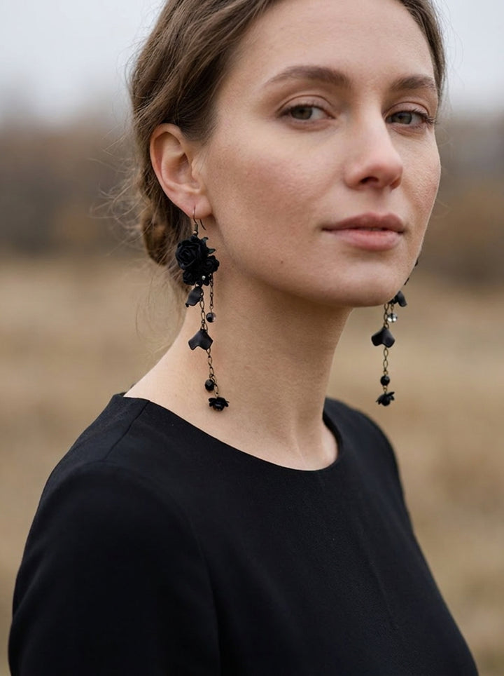 A woman wearing long black rose dangle earrings with small black flower charms and dark beads, photographed outdoors against a softly blurred tan field background.
