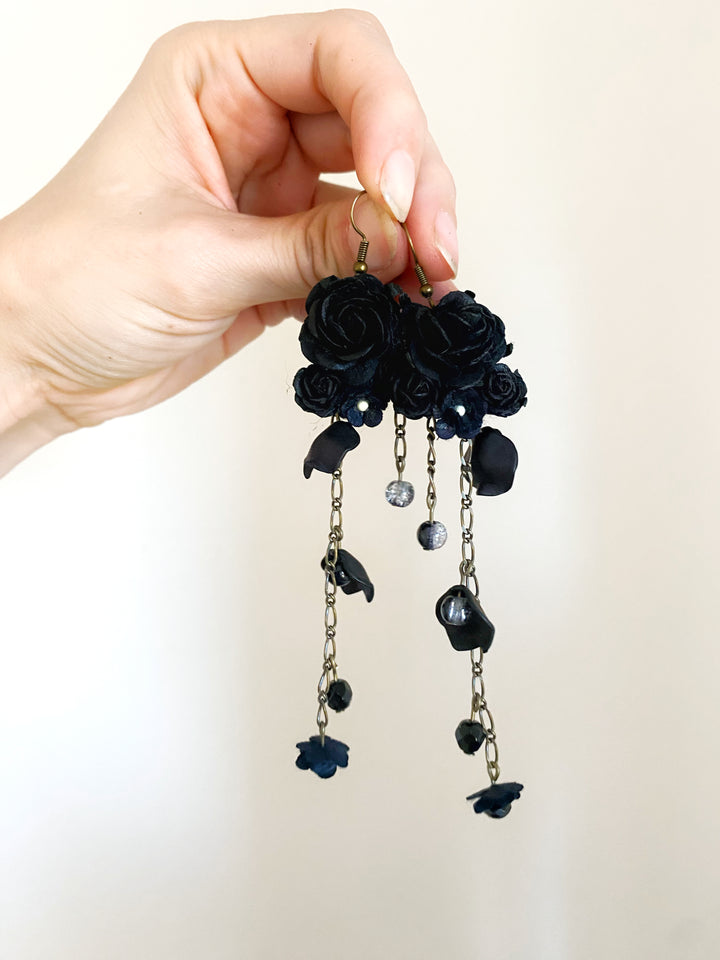 A hand holding a pair of long black floral dangle earrings with dark roses, small bead details, and dangling chain elements against a plain light background.