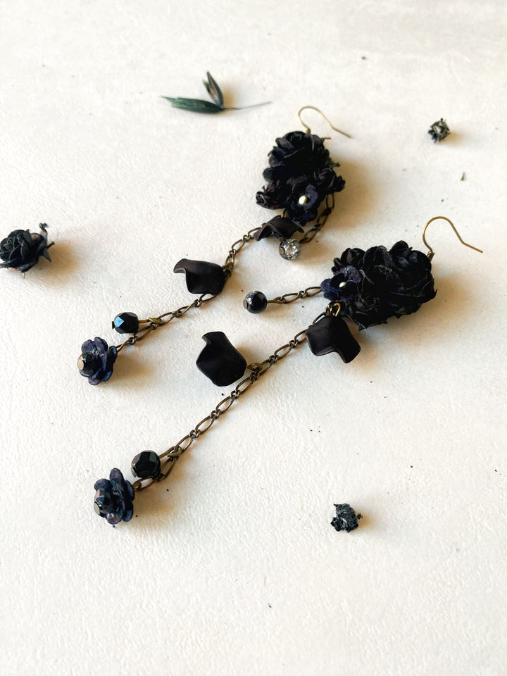 A pair of black floral dangle earrings with dark bronze chains and small black beads, displayed on a textured off-white surface with scattered dried flower petals.