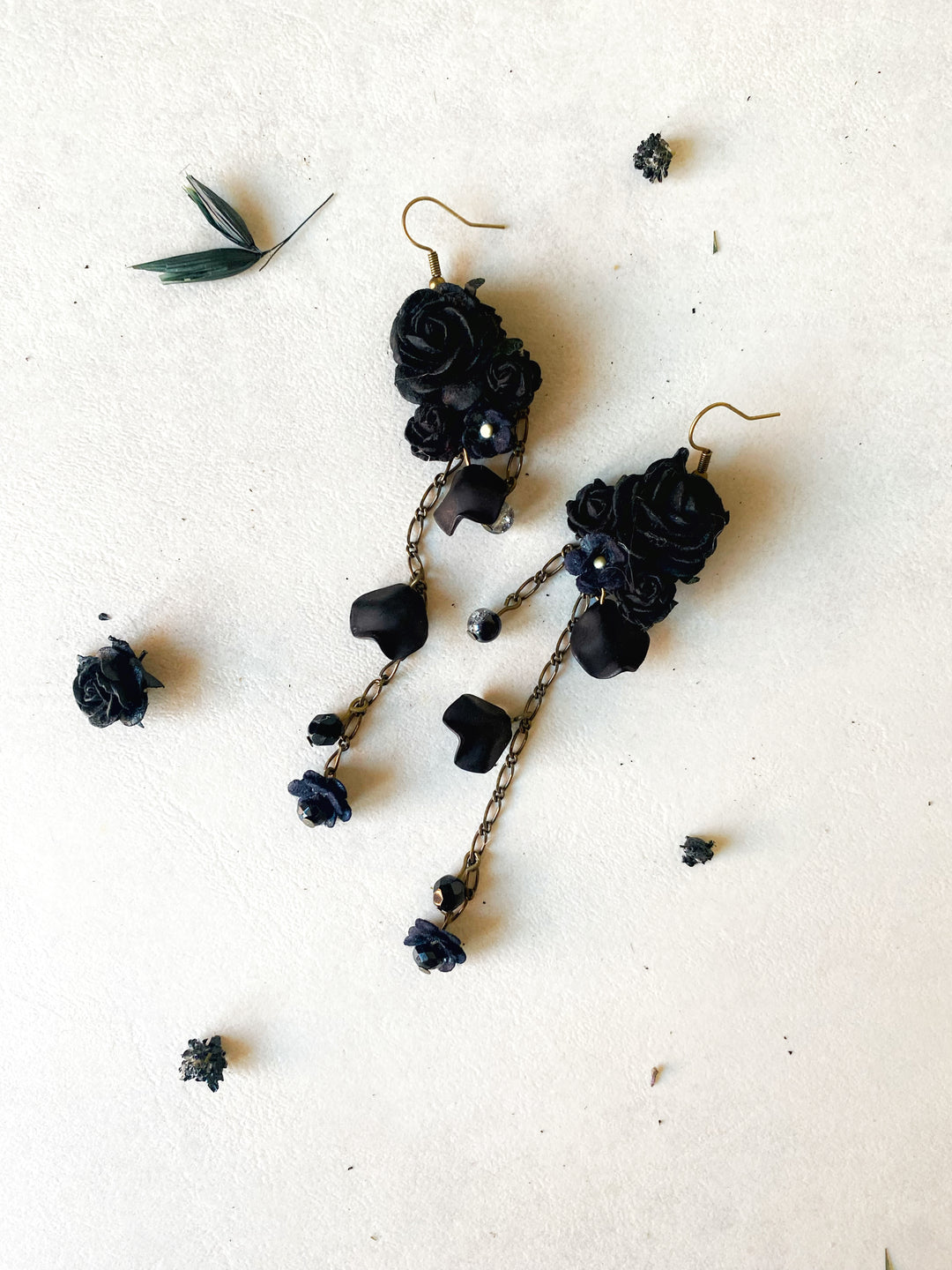 A pair of gothic dangle earrings featuring black fabric roses, dark beads, and twisted chains with small blue flower charms, laid flat on a textured off-white background with scattered dried flower petals.