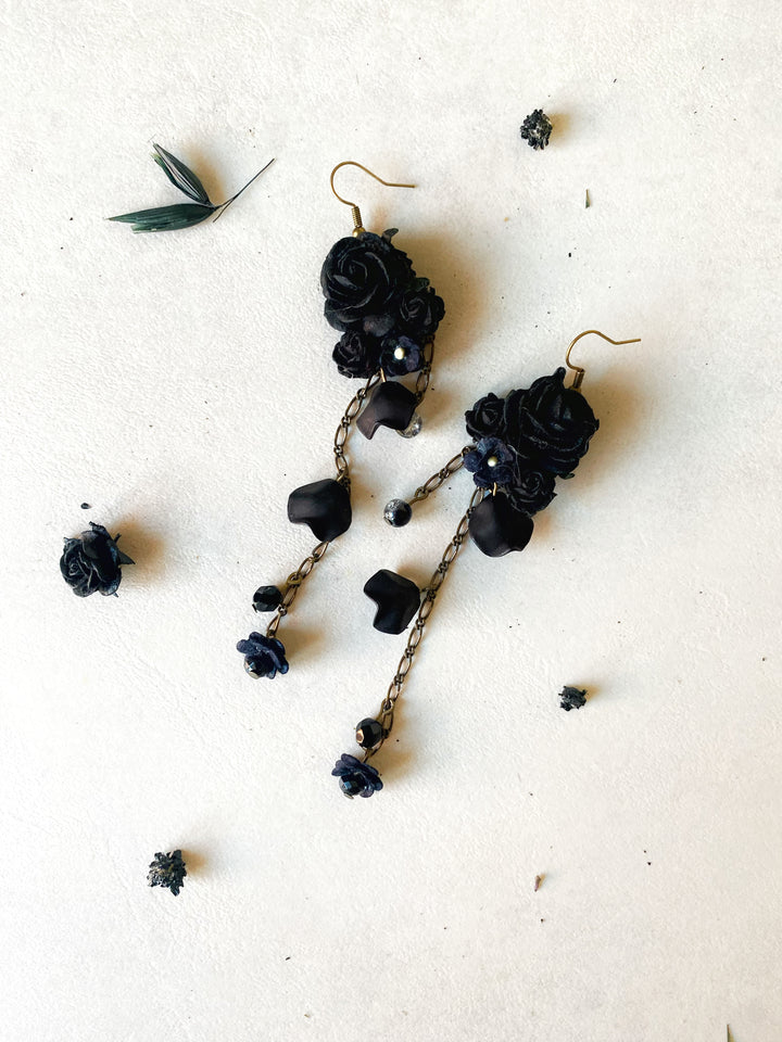 A pair of gothic dangle earrings featuring black fabric roses, dark beads, and twisted chains with small blue flower charms, laid flat on a textured off-white background with scattered dried flower petals.