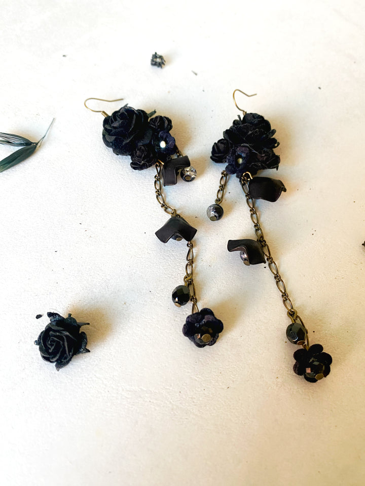 Black fabric rose drop earrings with antique gold-tone chains and beads, displayed on a light beige surface.