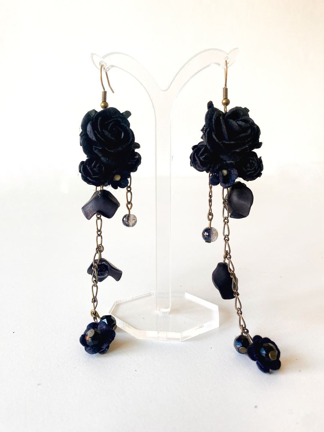 A pair of long black rose dangle earrings displayed on a clear acrylic stand against a white background. The earrings feature textured black fabric roses, dark metallic charms, and small dark beads on gold-tone hooks.
