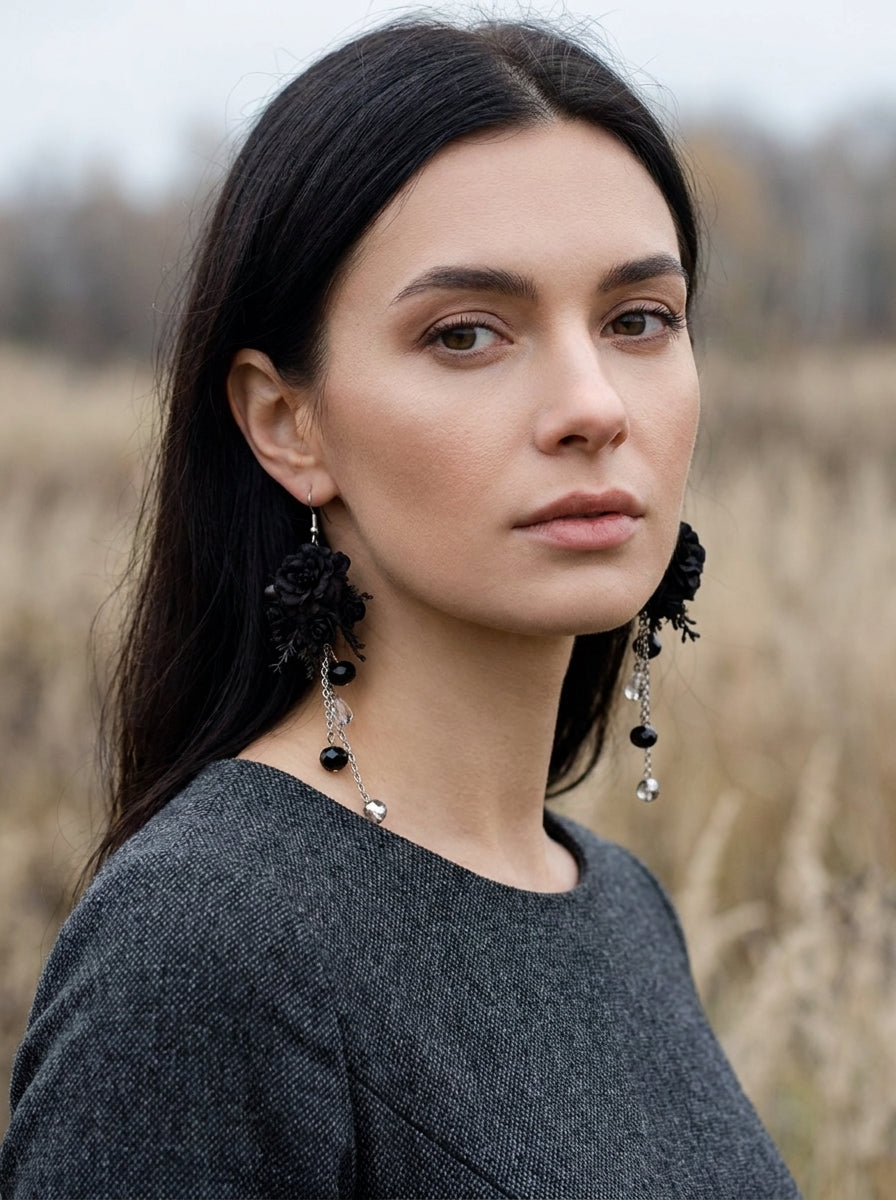 A woman with long dark hair wearing large black floral dangle earrings with silver-toned bead accents, photographed outdoors against a blurred field background.
