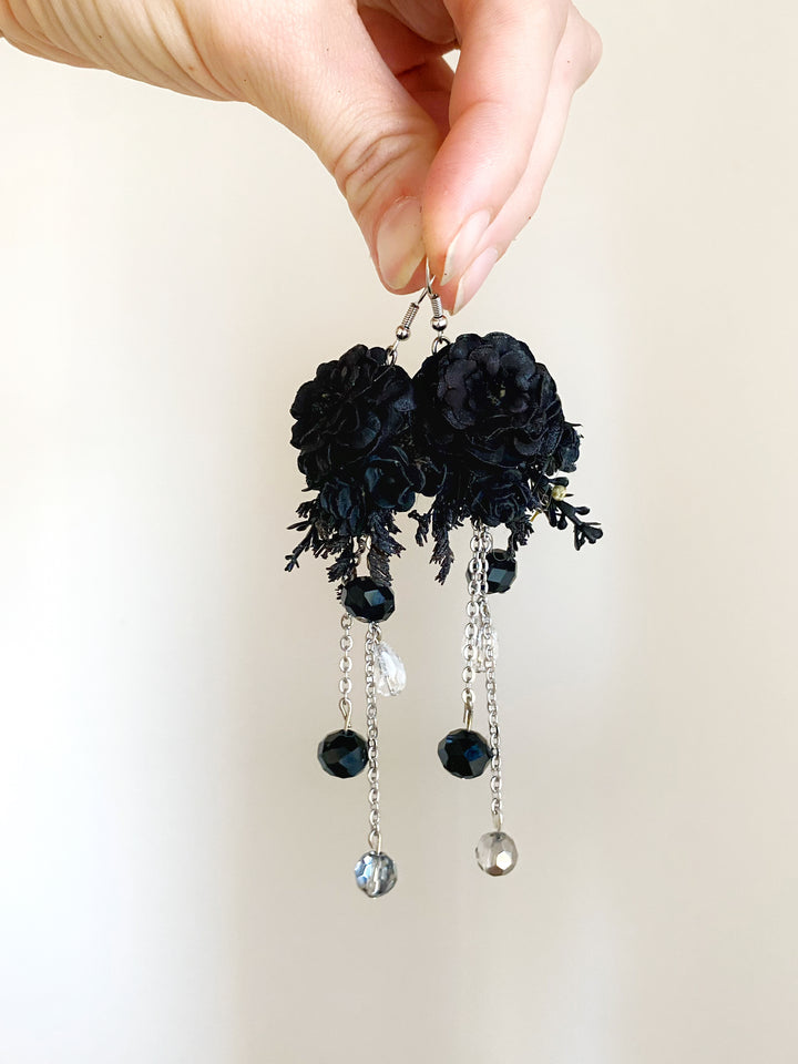 A pair of long black floral dangle earrings held by a hand against a plain light background. The earrings feature black fabric flowers, black lace details, and dangling black beads with silver-tone chains and clear crystal accents.