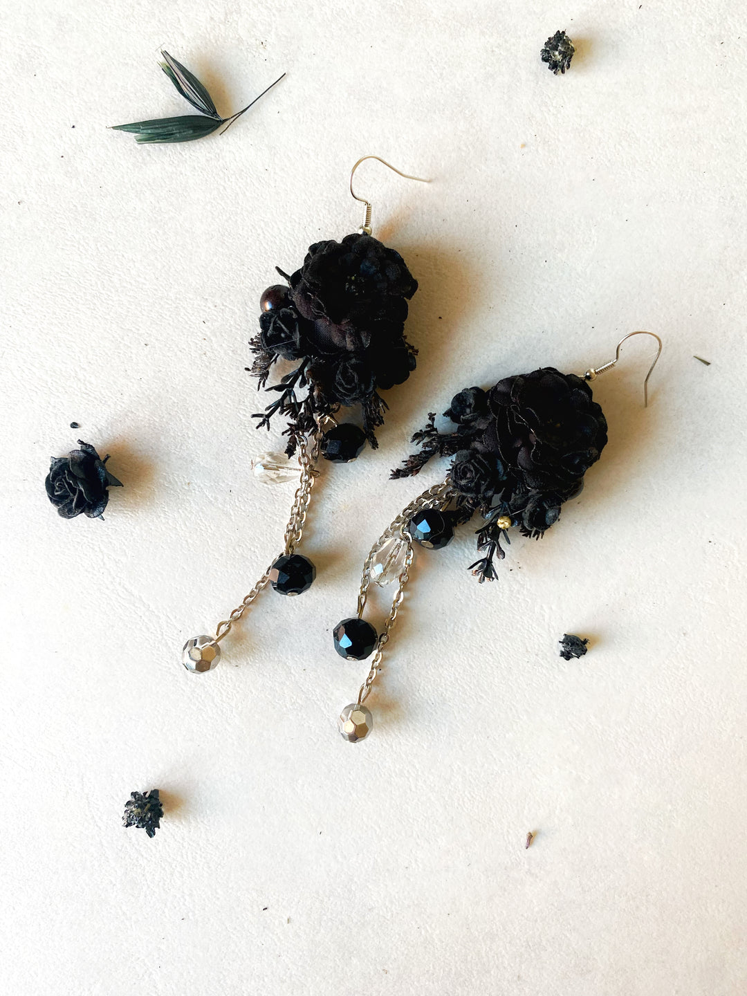 A pair of black floral dangle earrings with gold-tone chains, beads, and hooks, displayed on a textured off-white background with scattered dried botanical elements.