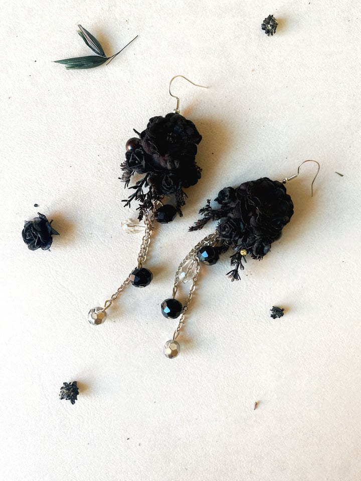 A pair of black floral dangle earrings with gold-tone chains, beads, and hooks, displayed on a textured off-white background with scattered dried botanical elements.