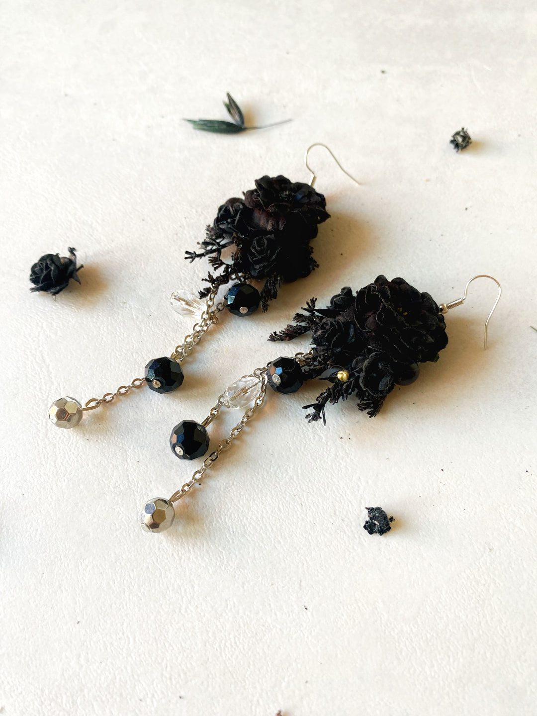 Black flower dangle earrings with silver-toned chains, faceted black beads, and clear crystal accents, displayed on a textured off-white surface with scattered dried botanicals.