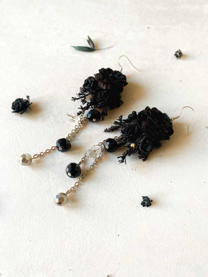 Black flower dangle earrings with silver-toned chains, faceted black beads, and clear crystal accents, displayed on a textured off-white surface with scattered dried botanicals.