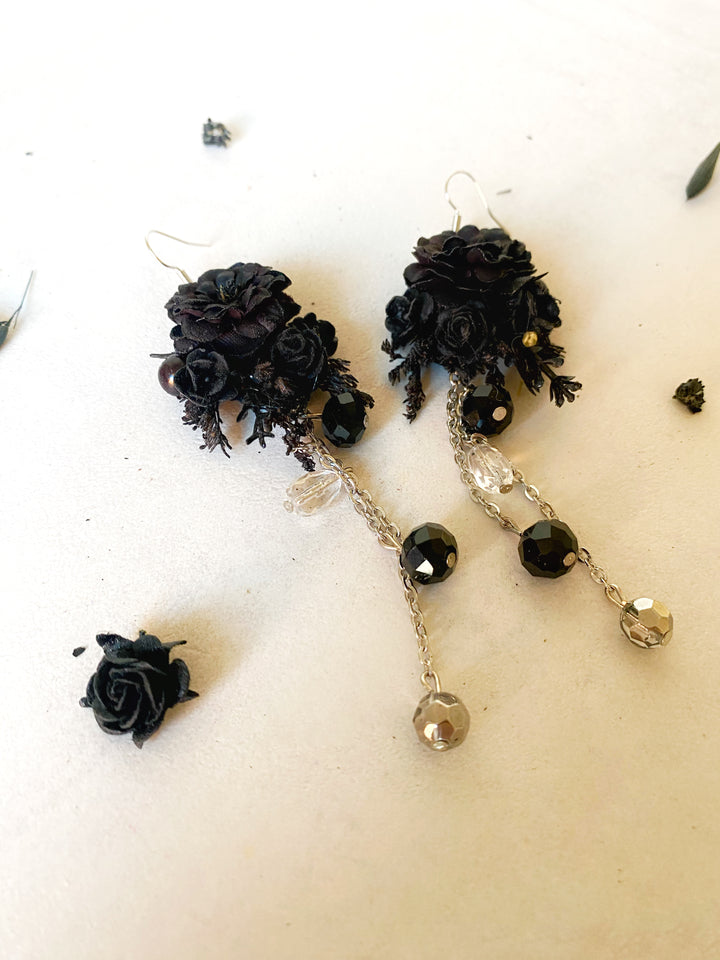 A pair of gothic-style dangle earrings featuring black fabric roses, faceted black beads, and clear crystal drops on silver chains, displayed on a light beige surface.