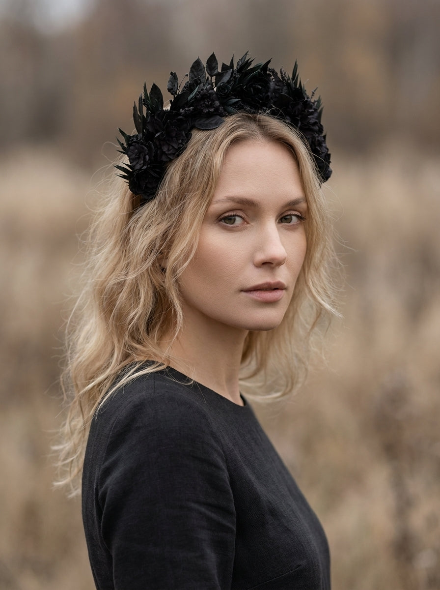 A woman with blonde hair wearing a black floral crown headband and a black long-sleeve top, photographed outdoors against a blurred natural background.