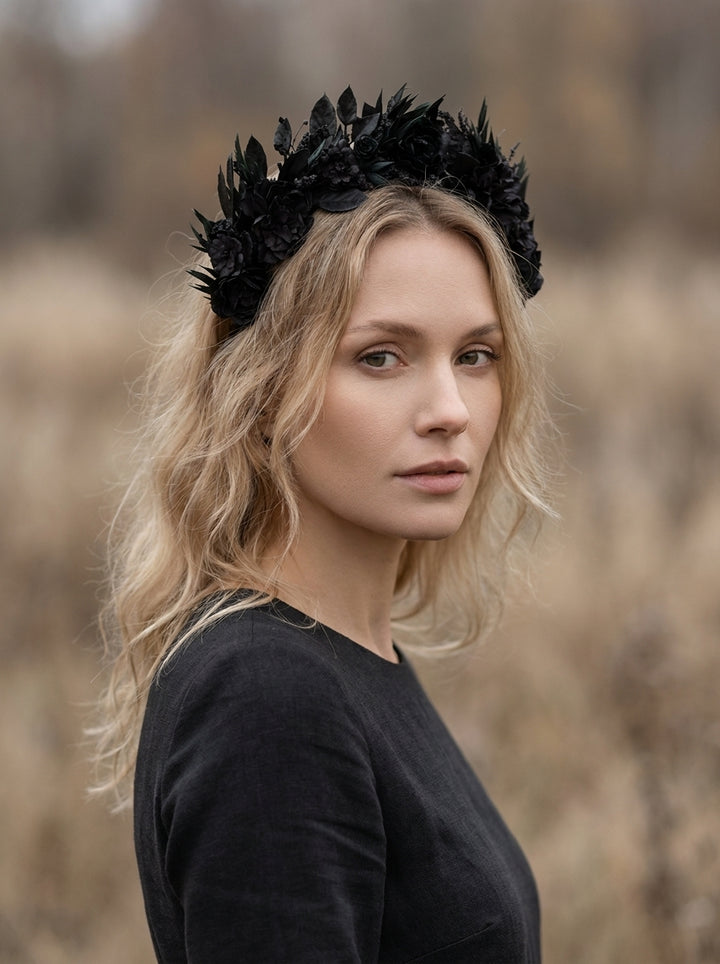 A woman with blonde hair wearing a black floral crown headband and a black long-sleeve top, photographed outdoors against a blurred natural background.