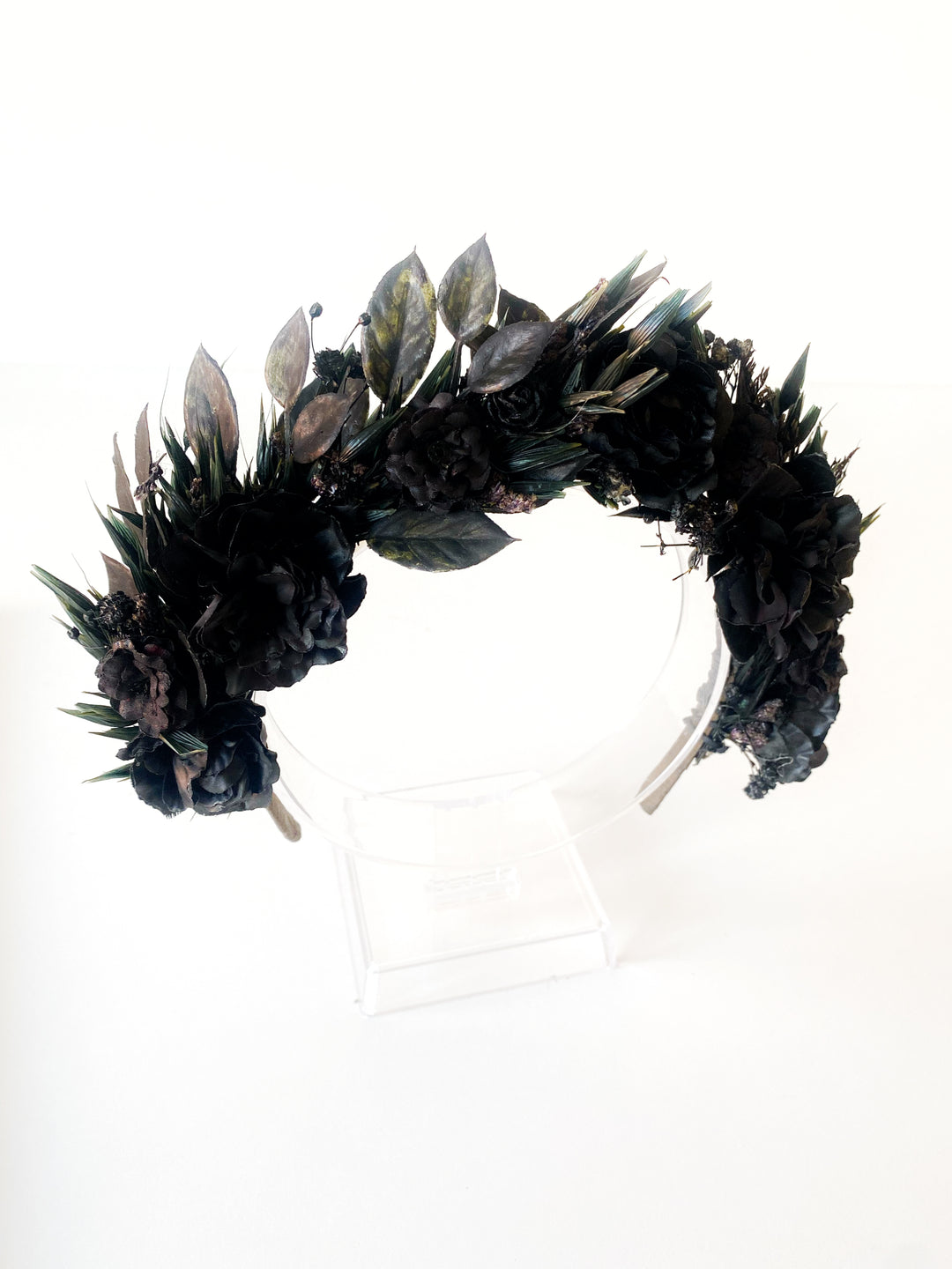 A black floral and leaf crown displayed on a clear acrylic stand against a plain white background.