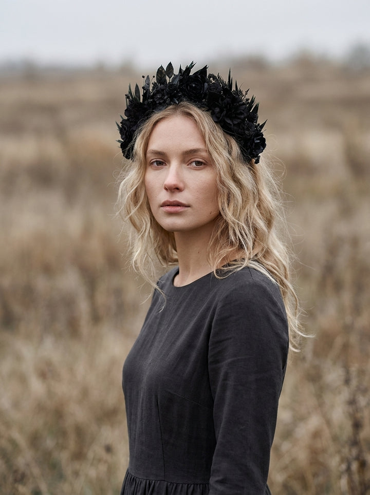 A blonde woman wearing a black leaf crown and a dark long-sleeve dress, standing in a dry grassy field.