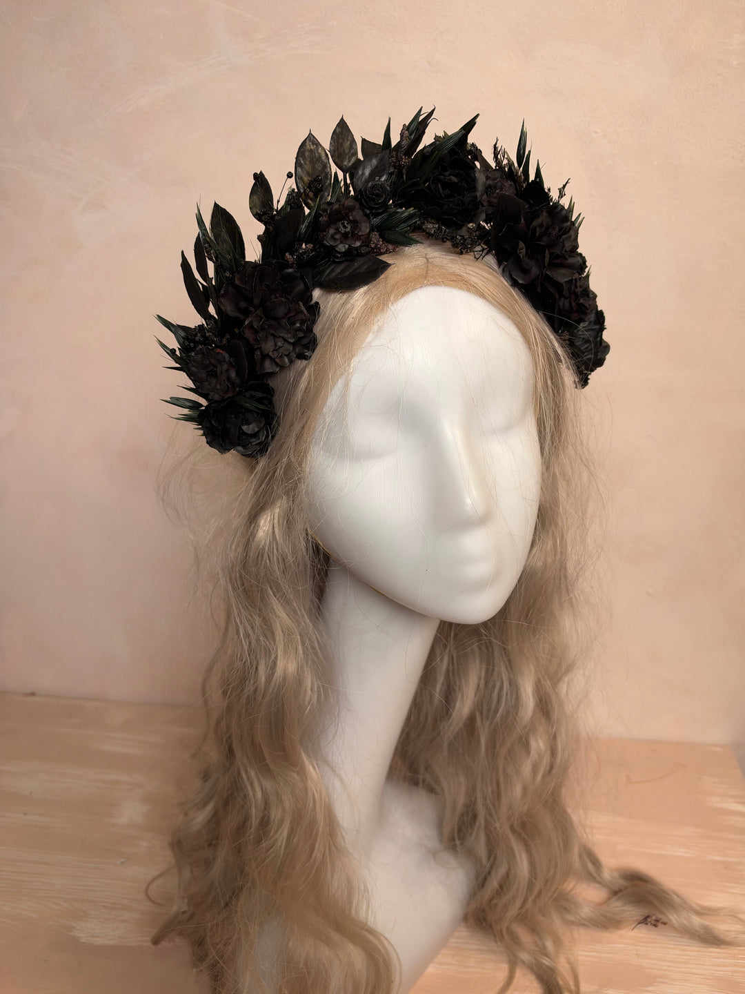 A black floral crown made of artificial dark flowers and leaves, displayed on a white mannequin head with long wavy blonde hair against a warm beige wall and light wood surface.