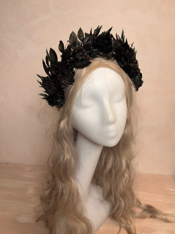 A black floral crown made of artificial dark flowers and leaves, displayed on a white mannequin head with long wavy blonde hair against a warm beige wall and light wood surface.
