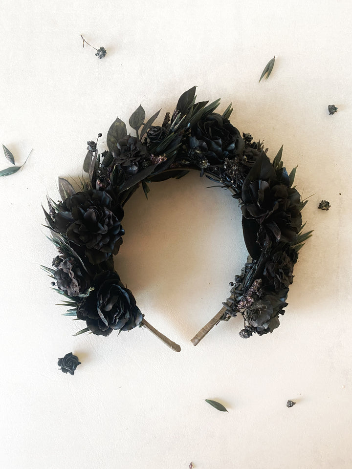 A black floral headband styled with black roses, dark green leaves, laid flat on a light beige background with scattered dried petals.