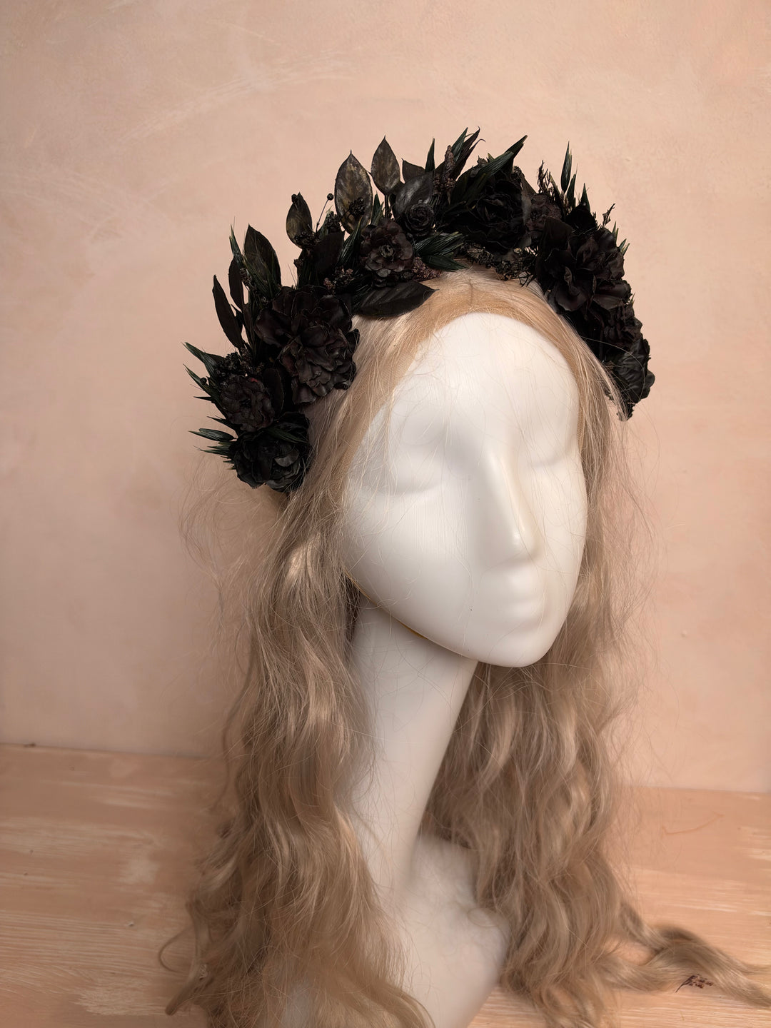 A black floral crown with dark leaves displayed on a white mannequin head with long wavy blonde hair, set against a warm beige textured wall.