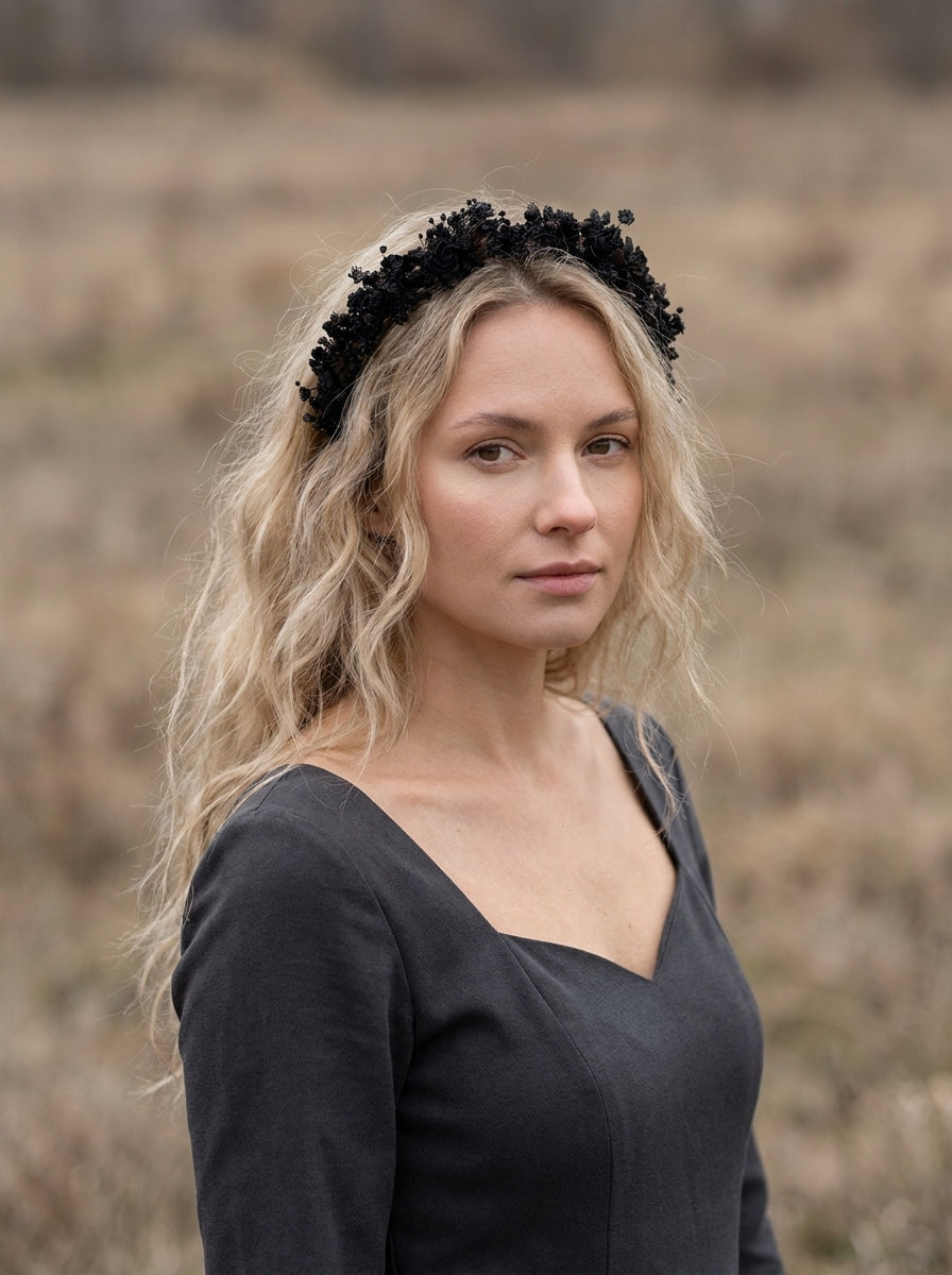 A woman with blonde hair wearing a black floral crown headband and a dark gray top, standing in a dry, grassy field.