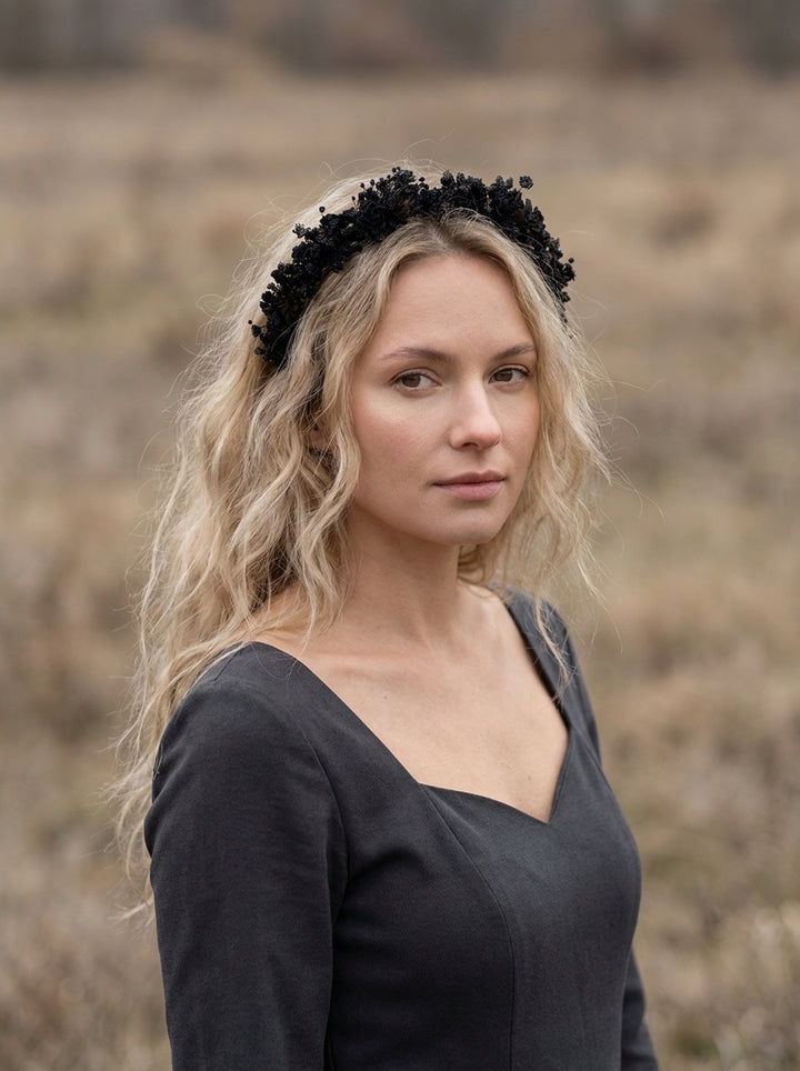 A woman with blonde hair wearing a black floral crown headband and a dark gray top, standing in a dry, grassy field.