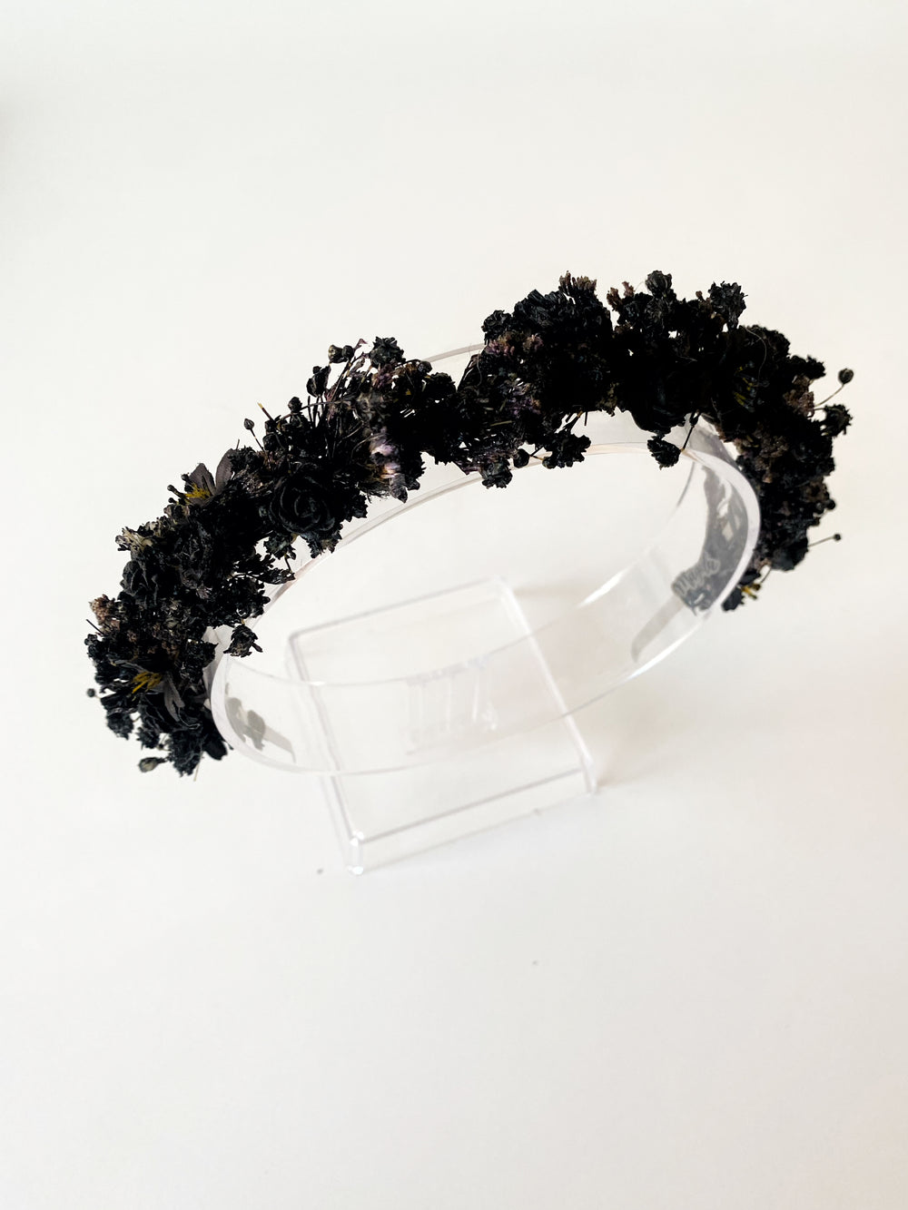 A black floral crown made of dried and artificial black flowers, displayed on a clear acrylic stand against a plain white background.