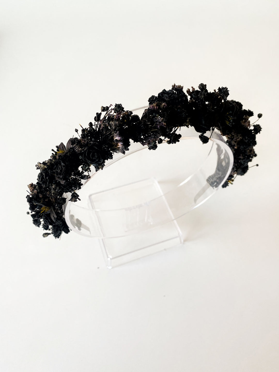 A black floral crown made of dried and artificial black flowers, displayed on a clear acrylic stand against a plain white background.