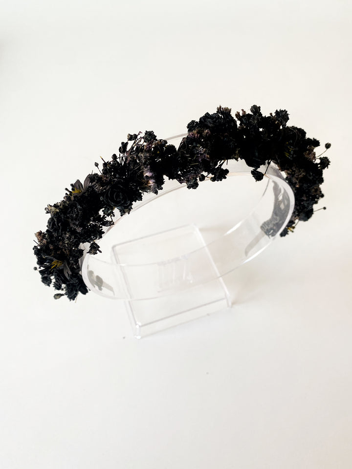 A black floral crown made of dried and artificial black flowers, displayed on a clear acrylic stand against a plain white background.