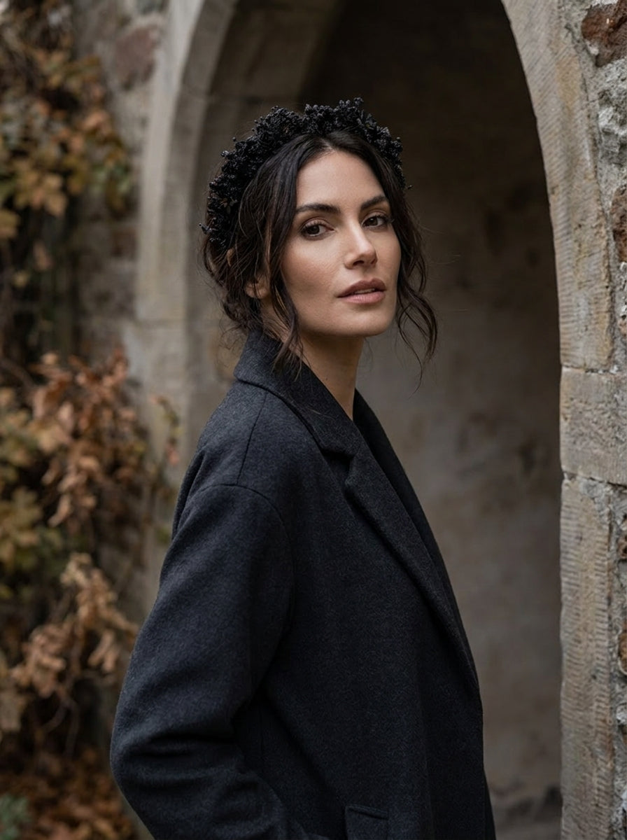 A woman wearing a black floral headpiece and a dark charcoal gray blazer, standing in a stone archway with autumn foliage on the left.
