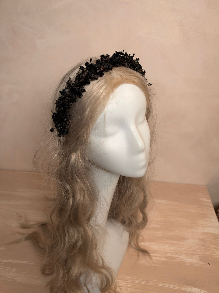 A black floral lace headband displayed on a blonde mannequin head with long wavy hair, set against a beige wall and light wood surface.