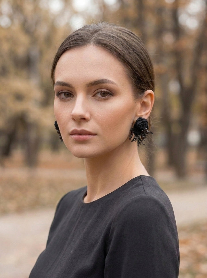 A woman with her hair pulled back wears large, dramatic black flower-shaped earrings and a black top, photographed outdoors against a blurred autumn park background.