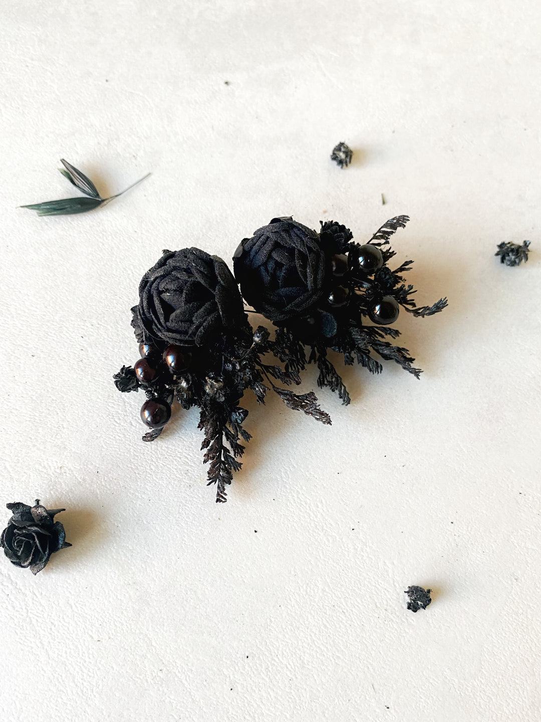 A pair of black fabric peony earrings with  displayed on a textured off-white surface with scattered decorative black rose petals.