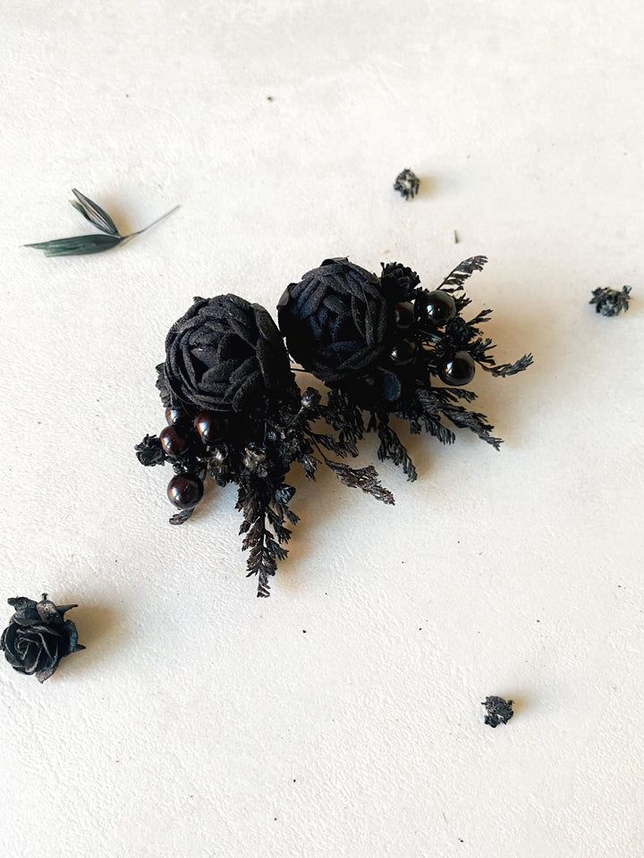 A pair of black fabric peony earrings with  displayed on a textured off-white surface with scattered decorative black rose petals.