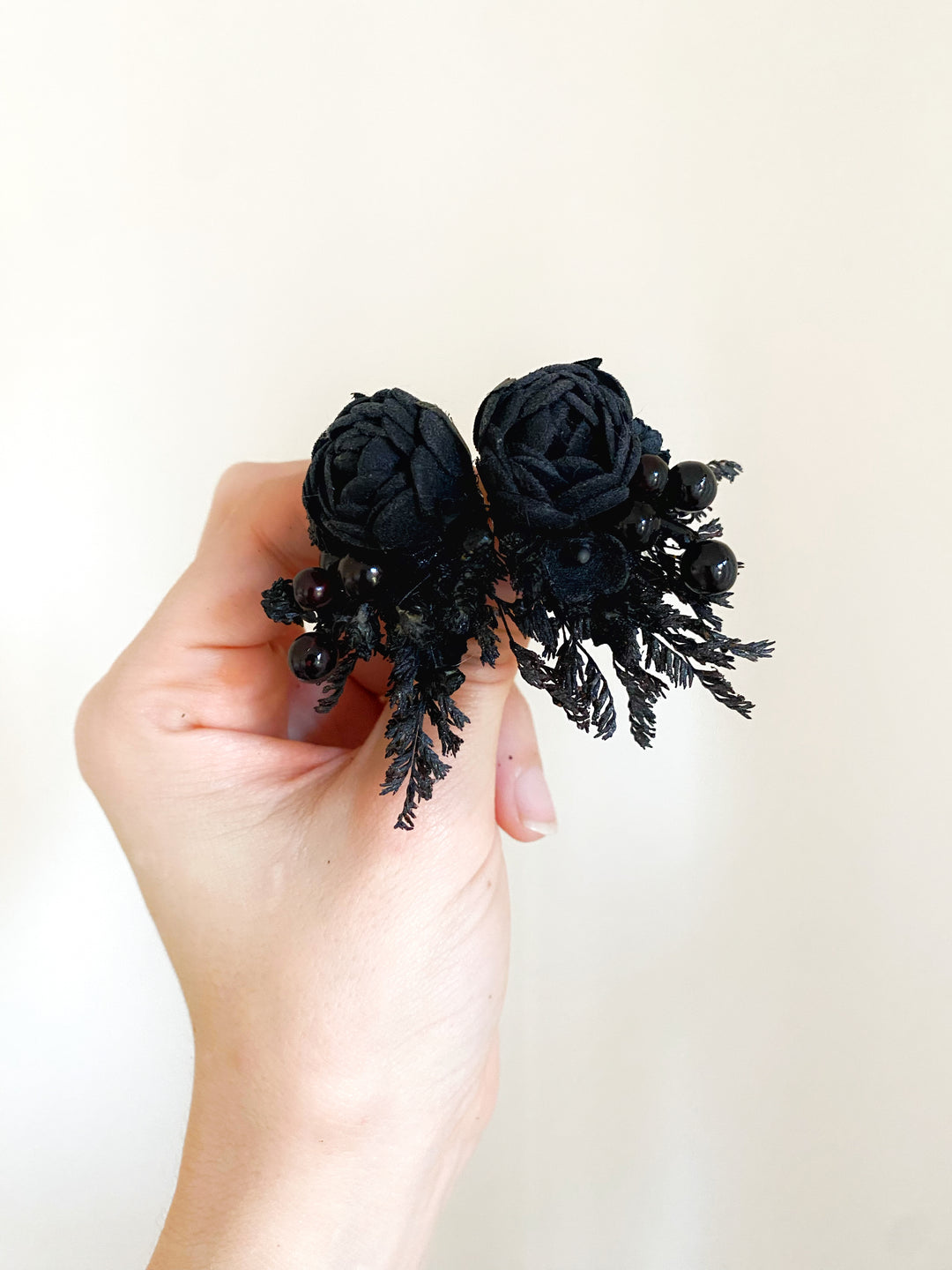 A hand holding a black floral earrings  with two fabric peonies, black berries, and textured black leafy accents against a plain off-white background.
