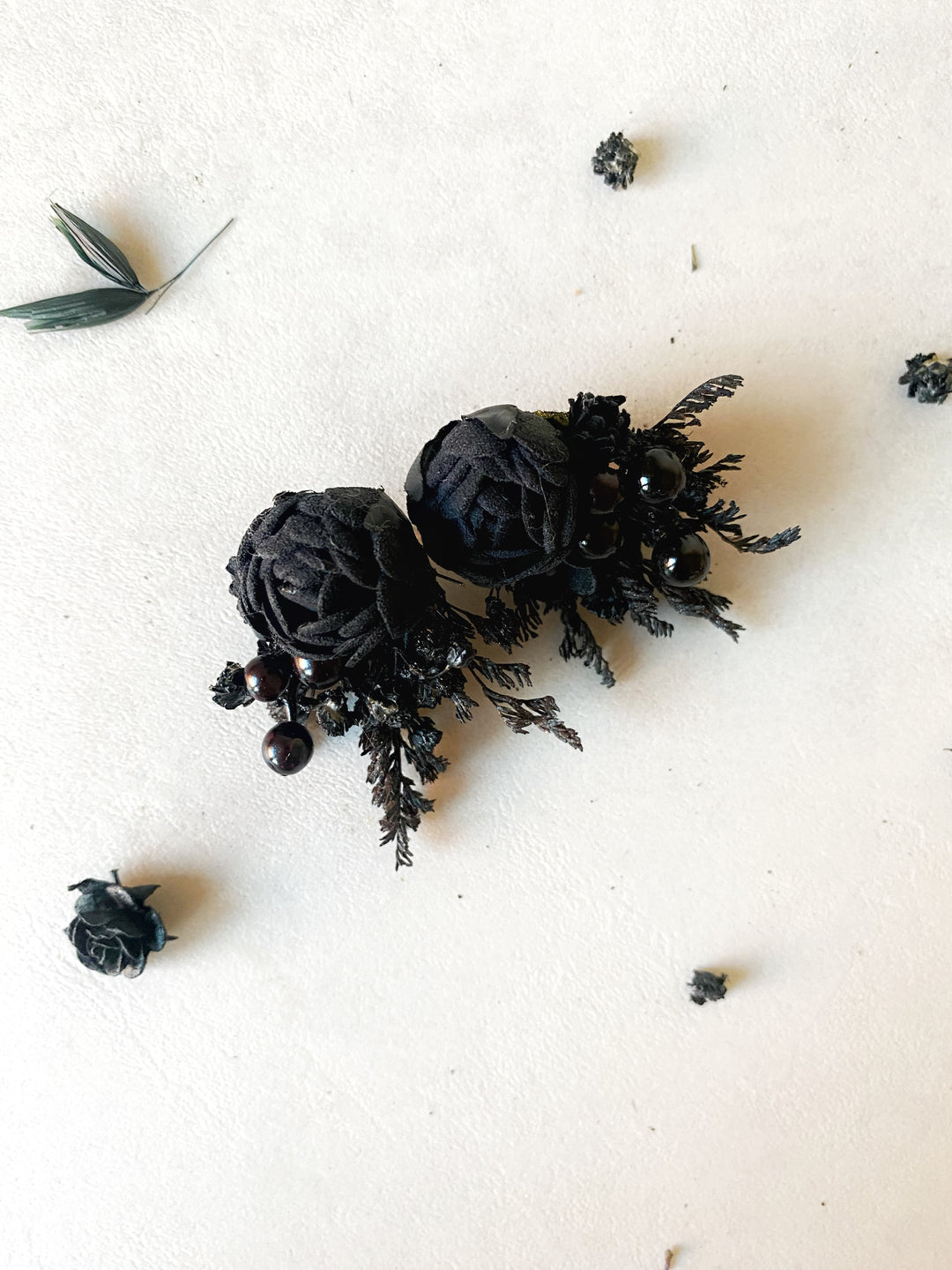 A pair of black fabric rose stud earrings with dark green pineconelike accents and small black bead details, displayed on a textured off-white background with scattered dried botanical elements.